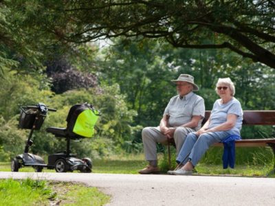 an elderly couple sits on a bench near an EV scooter powered by a charging station