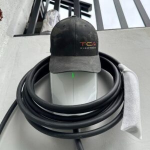 A Tesla charging connector with a coiled black cable, topped with a black cap featuring the "TCA ELECTRIC" logo